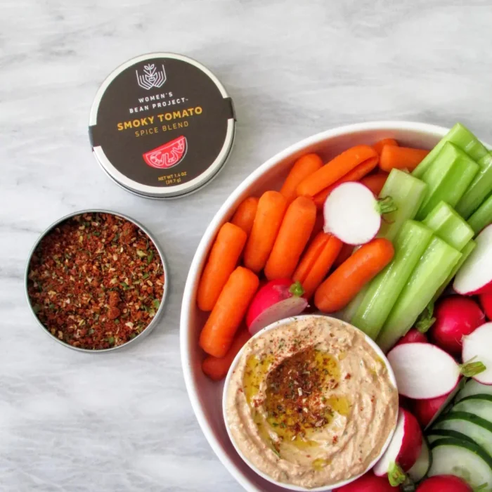 An open tin of Women's Bean Project Smoky Tomato Spice Blend next to a plate of vegetables and smoky tomato hummus.